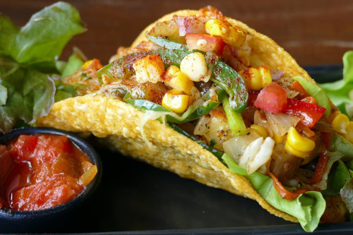 Crispy corn tortilla taco filled with corn, peppers and tomato, with a bowl of red salsa