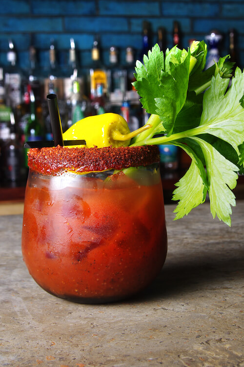 Bloody Mary with a chili-salt rim, celery and pickled yellow pepper before a liquor shelf