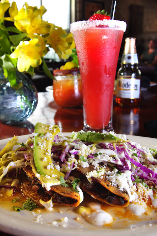 Two tacos with crumbled cheese, purple cabbage and avocado beside a strawberry drink and Modelo bottle