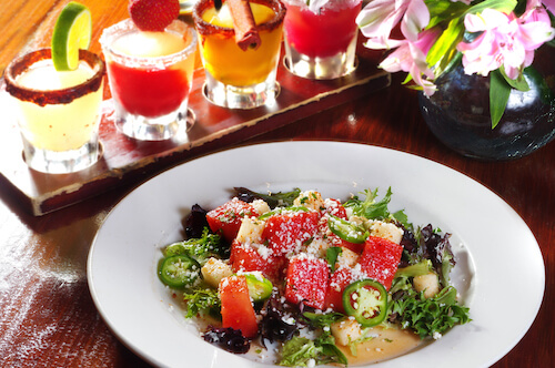 Watermelon salad on a white plate with four colorful rimmed drinks on a tray behind