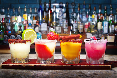 Flight of four rimmed margaritas on a wooden board before a wall of liquor bottles
