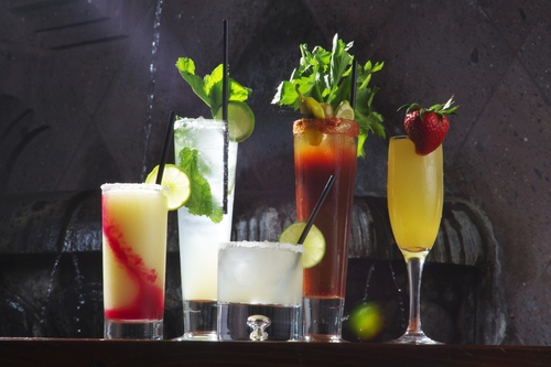 Five cocktails in different glasses with lime, mint and a strawberry garnish against a dark wall