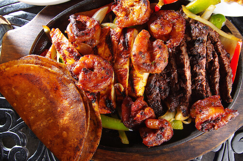 Sizzling fajita skillet with grilled shrimp, sliced steak, chicken and peppers beside a folded tortilla