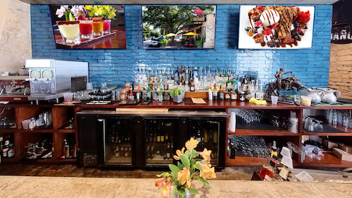 Bar with a blue tile wall, three wall-mounted screens, a liquor shelf and a built-in cooler
