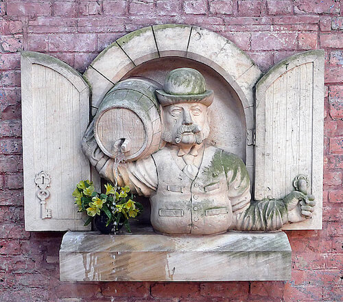 Stone relief of a bearded man in a hat holding a barrel, set in brick wall